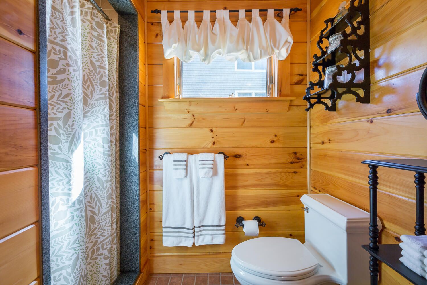 A wooden cabin bathroom with a toilet, two towels, a window with a curtain, and a wooden shelf with a candle.