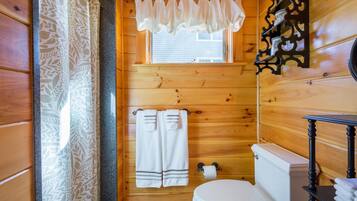 A wooden cabin bathroom with a toilet, two towels, a window with a curtain, and a wooden shelf with a candle.