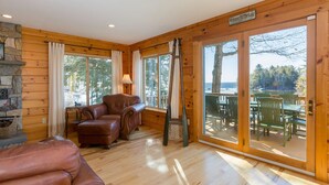 A cabin interior with a stone fireplace, leather sofa, and a large sliding glass door leading to an outdoor deck with tables and chairs. - Maple Cove Lodge on Lake Sebago Waterfront! (Casco)