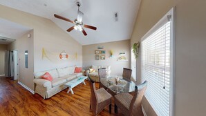 A spacious living room with a wooden floor, a glass dining table, wicker chairs, a ceiling fan, and a large window with blinds. - Cute beach house, 2 min walk to the beach (Myrtle Beach)