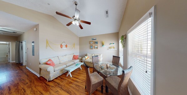 A spacious living room with a wooden floor, a glass dining table, wicker chairs, a ceiling fan, and a large window with blinds.