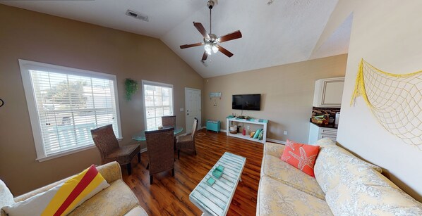 A living room with a ceiling fan, a dining area with wicker chairs, a sofa with a starfish pillow, and a TV on the wall.