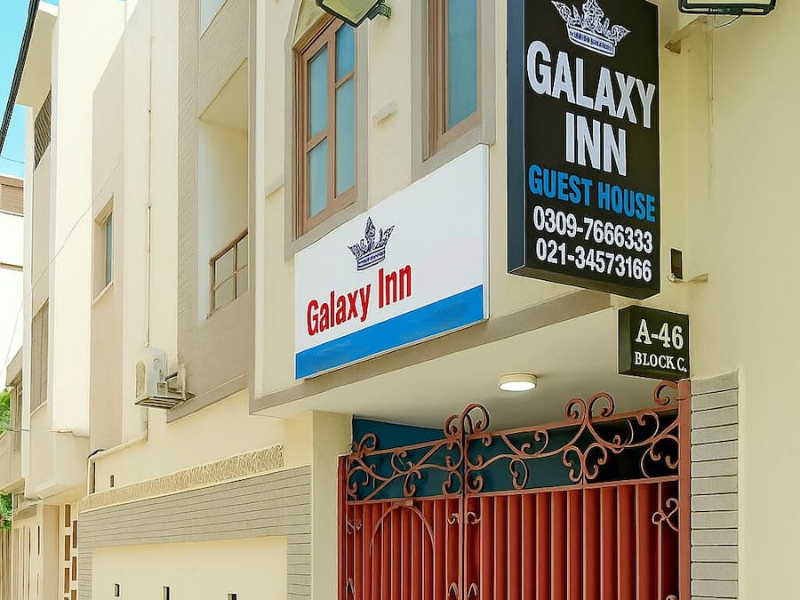 A Galaxy Inn guest house with a large red gate, beige walls, and a signboard displaying the name and contact information.