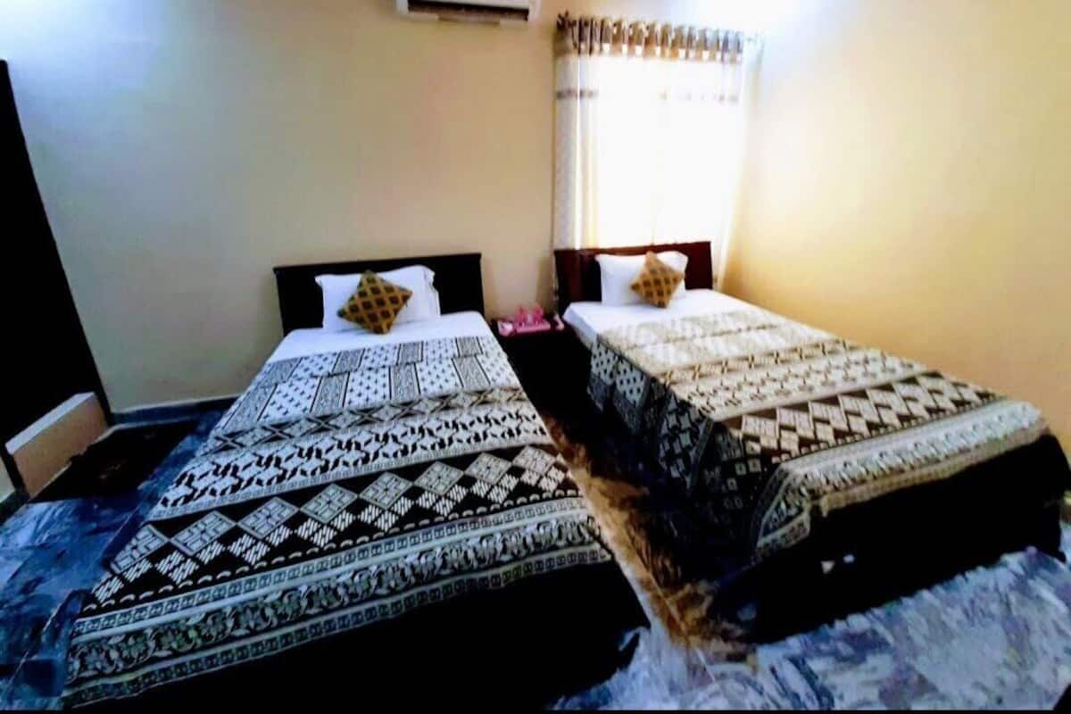 A room with two beds, each with patterned bedding, a window with curtains, and an air conditioning unit on the wall.