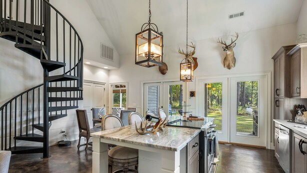 A modern kitchen with a central island, a black spiral staircase, and a deer head mounted on the wall. - Woodland Gameday Getaway Near Tuscaloosa – Pond Views, Pavilion, Firepits & More (Akron)