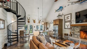 A spacious living area with a spiral staircase, a fireplace, and various decorative antlers. - Woodland Gameday Getaway Near Tuscaloosa – Pond Views, Pavilion, Firepits & More (Akron)