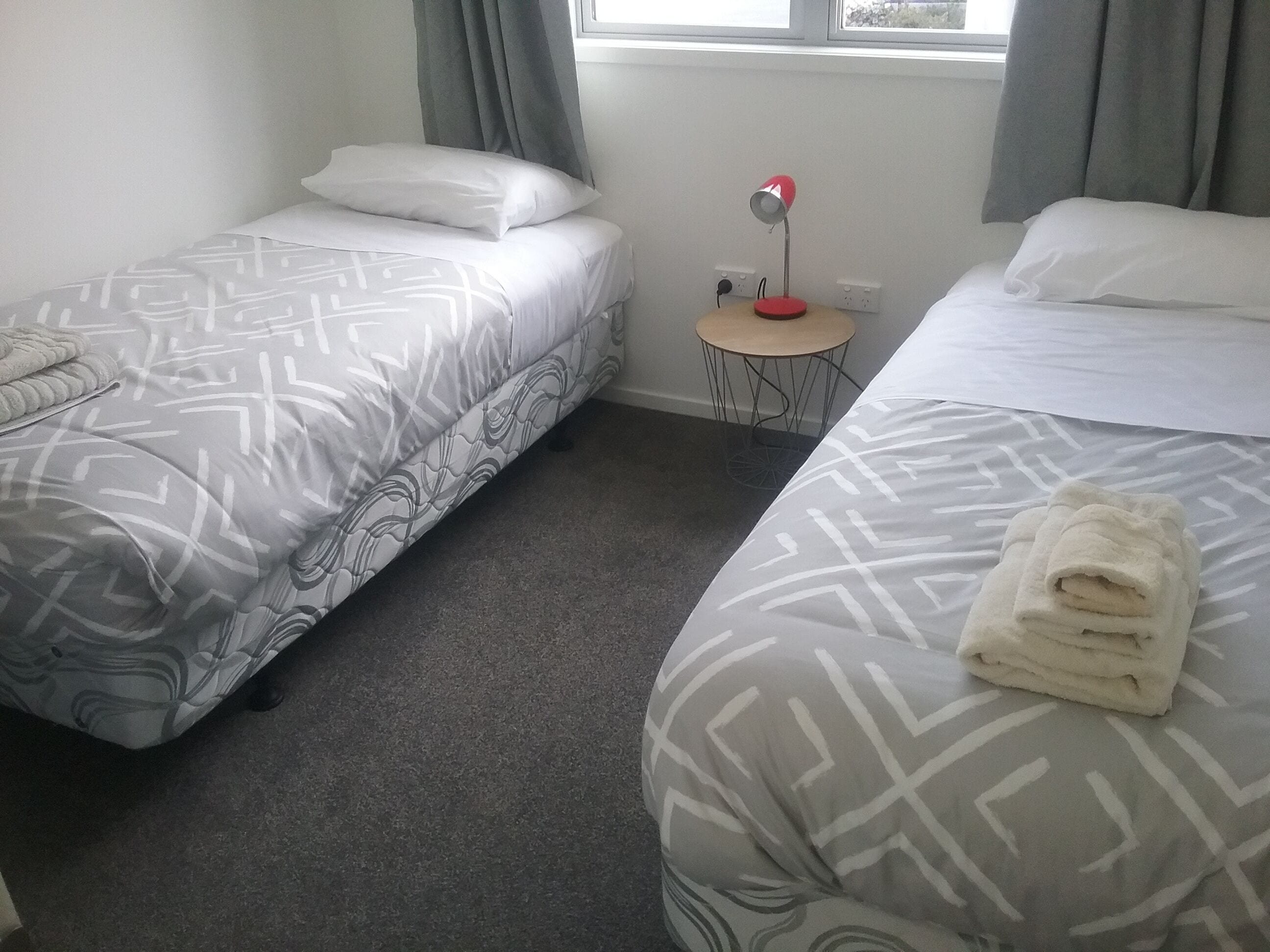 A hotel room with two beds, a small table with a lamp, and a window with curtains.