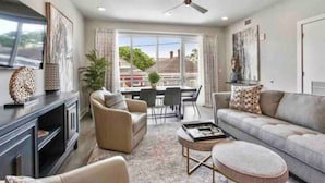 A modern living room with a sofa, armchair, coffee table, and TV stand. There is a dining area with a table and chairs, and a large window providing a view of the outside. - New Luxurious & Beautiful! - 2br/2ba w/ Pool! (New Orleans)