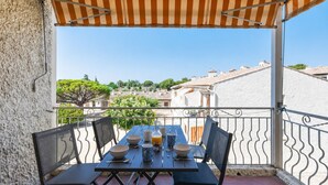 A balcony with a table set for two, chairs, a striped awning, and a view of buildings and trees. - Appartement Provenza - Welkeys (Saint-Cyr-sur-Mer)