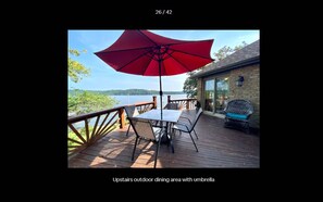An outdoor dining area with a red umbrella, a table, and chairs on a wooden deck overlooking a body of water.