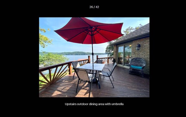 Un área de comedor al aire libre con una sombrilla roja, una mesa y sillas en una terraza de madera con vistas al agua.