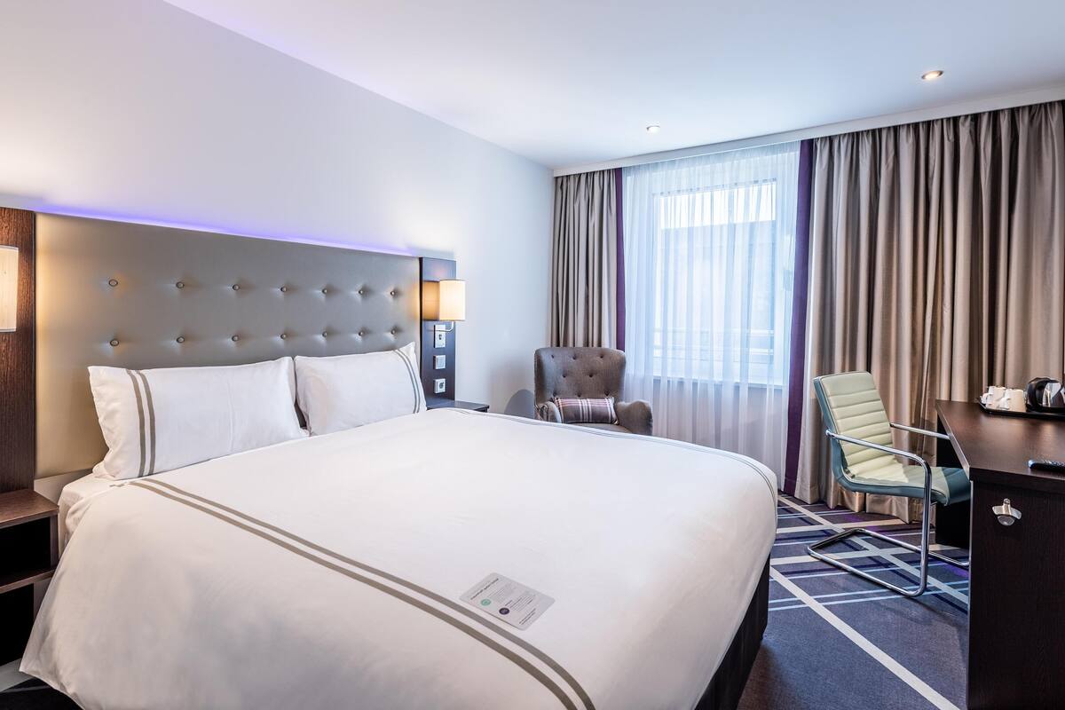 A hotel room with a large bed, a chair, a desk, and a window with curtains.