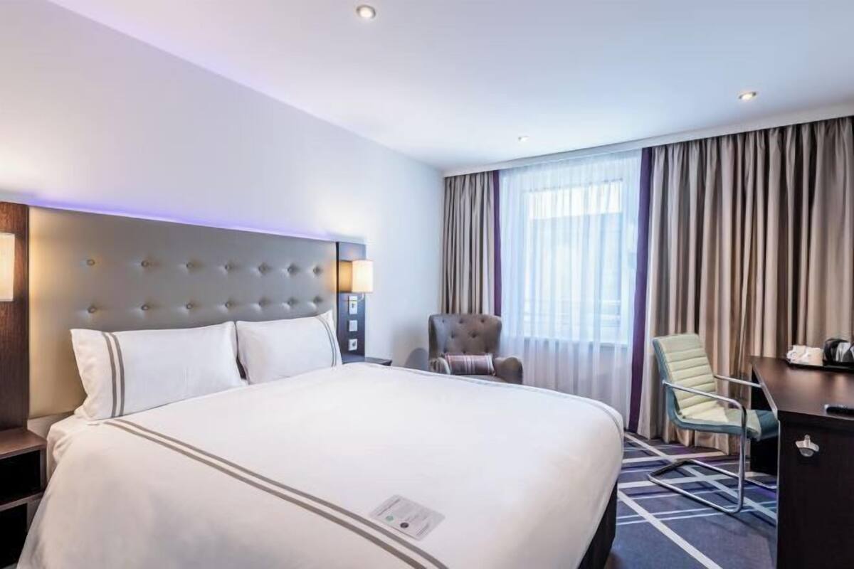 A hotel room with a large bed, a headboard with lights, a desk, a chair, and a window with curtains.