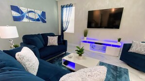 Un salon moderne avec un canapé bleu, une table basse blanche agrémentée d’une plante et un téléviseur à écran plat fixé au mur.