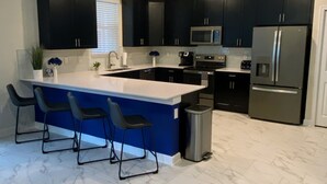 Une cuisine moderne avec des meubles de couleur sombre, un réfrigérateur en acier inoxydable, un four à micro-ondes et un évier. On y trouve trois tabourets de bar et une poubelle.
