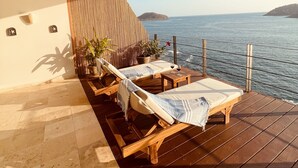 Une terrasse en bois avec des chaises longues et une table, donnant sur une étendue d’eau avec une île visible au loin.