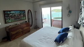 Une chambre avec un grand lit, un ventilateur de plafond, une commode, un tableau et une vue sur l’océan par une porte coulissante.