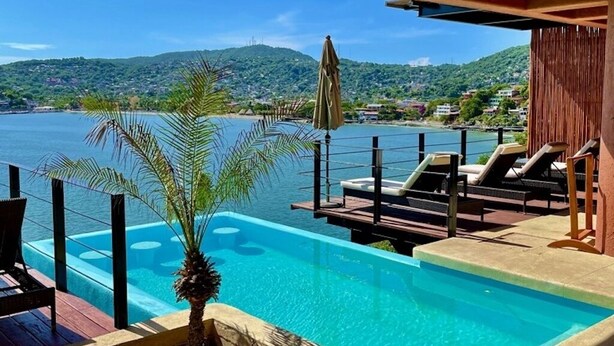 Un espace piscine aménagé avec des chaises longues et un palmier, offrant une vue sur une étendue d’eau et des collines.