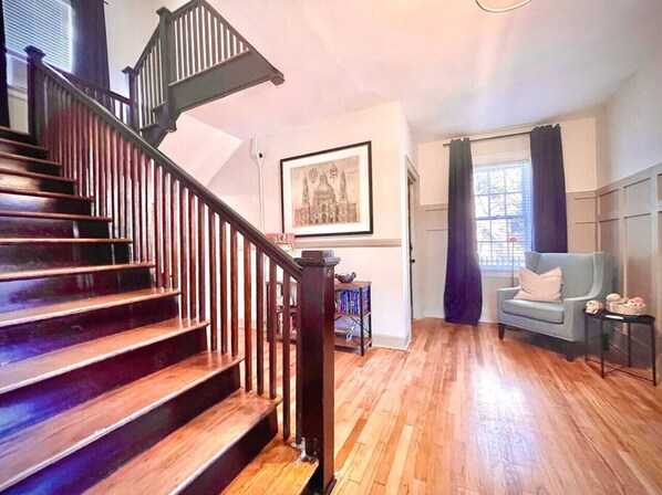 Una escalera con escalones de madera y barandilla negra, un cuadro en la pared, una ventana con cortinas y un sillón azul.