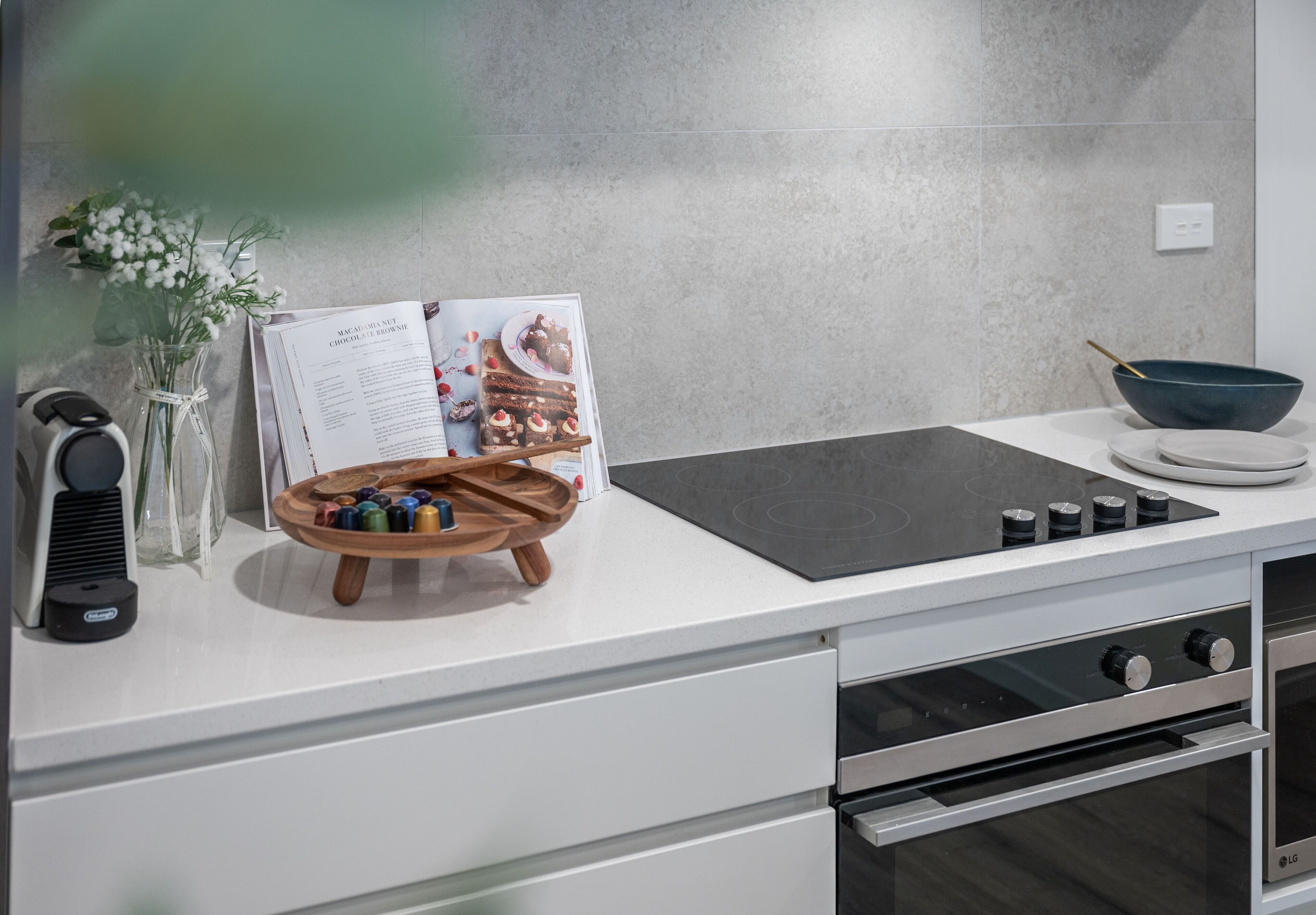 Una cocina con encimera blanca, placa de inducción negra, estante de madera para especias, un florero con flores y un libro de cocina.