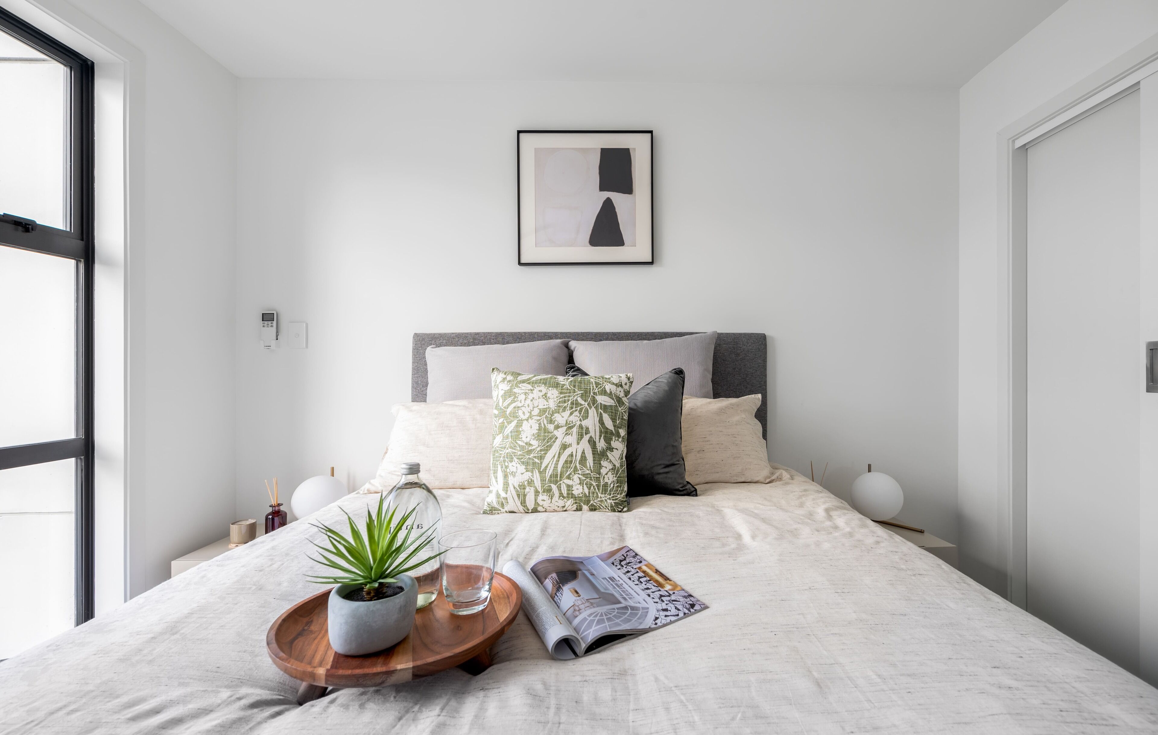 A modern bedroom with a bed, a bedside table with a plant, a magazine, and a framed picture on the wall.