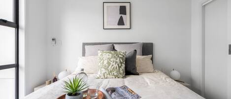 A modern bedroom with a bed, a bedside table with a plant, a magazine, and a framed picture on the wall.