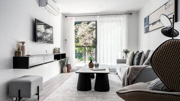 A modern living room with a sofa, a coffee table, a side table, a TV mounted on the wall, and a large window with curtains.