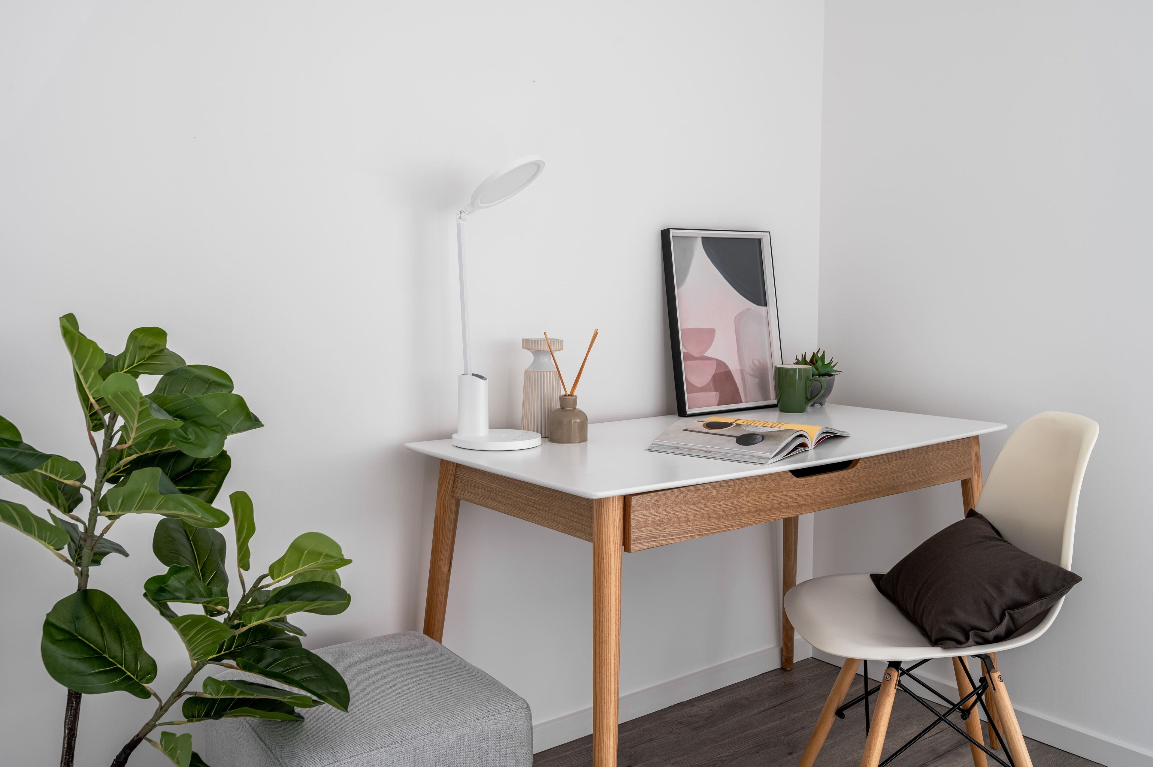 A minimalist workspace with a white desk, a modern lamp, a framed abstract painting, and a potted plant.