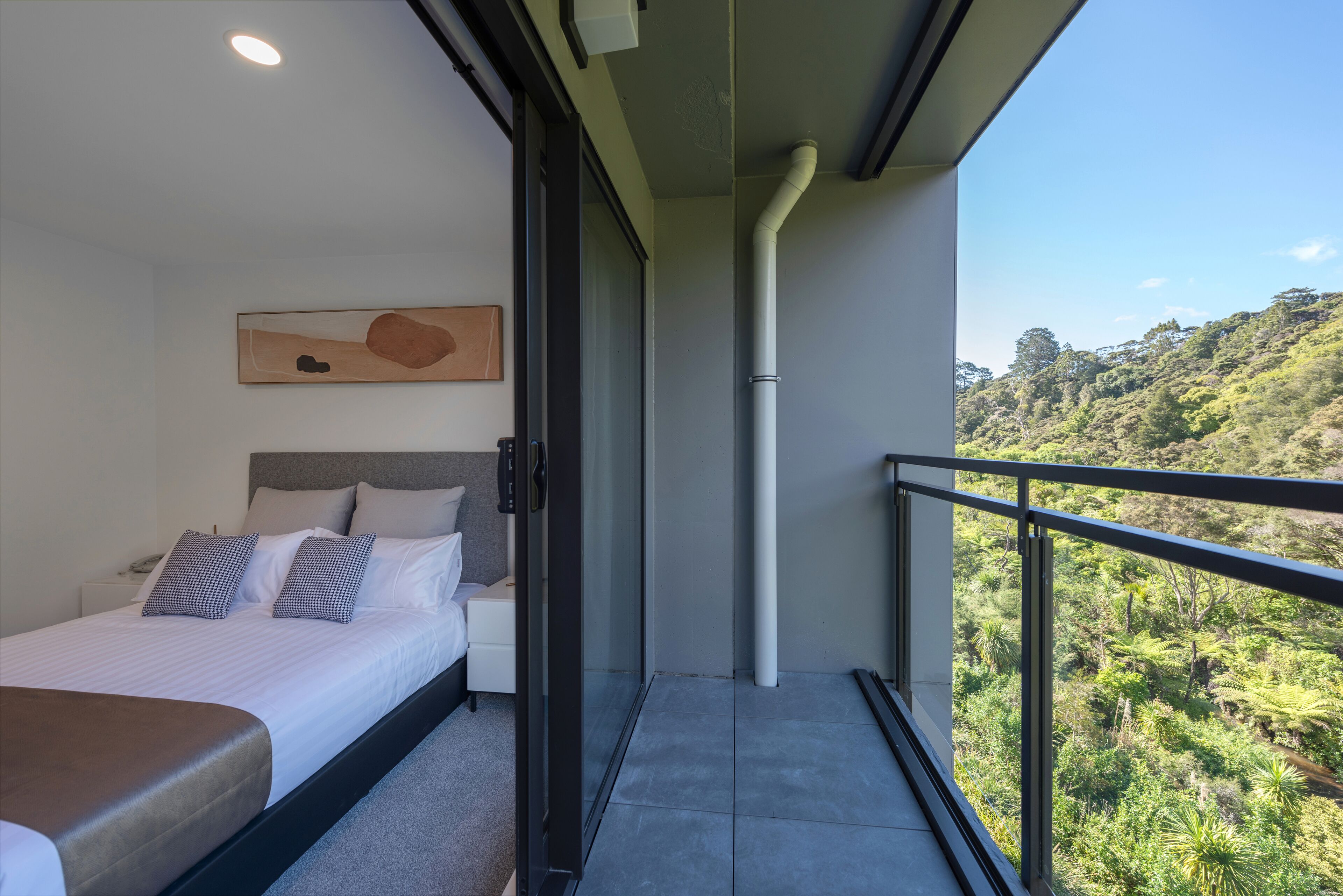 A modern bedroom with a bed, a bedside table, a painting on the wall, and a balcony with a view of a lush landscape.