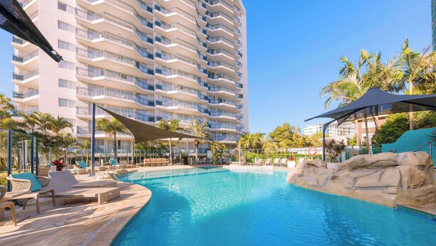 A hotel pool area with lounge chairs and umbrellas, surrounded by palm trees and a tall building. - Club Wyndham Kirra Beach, Trademark Collection by Wyndham (Coolangatta)