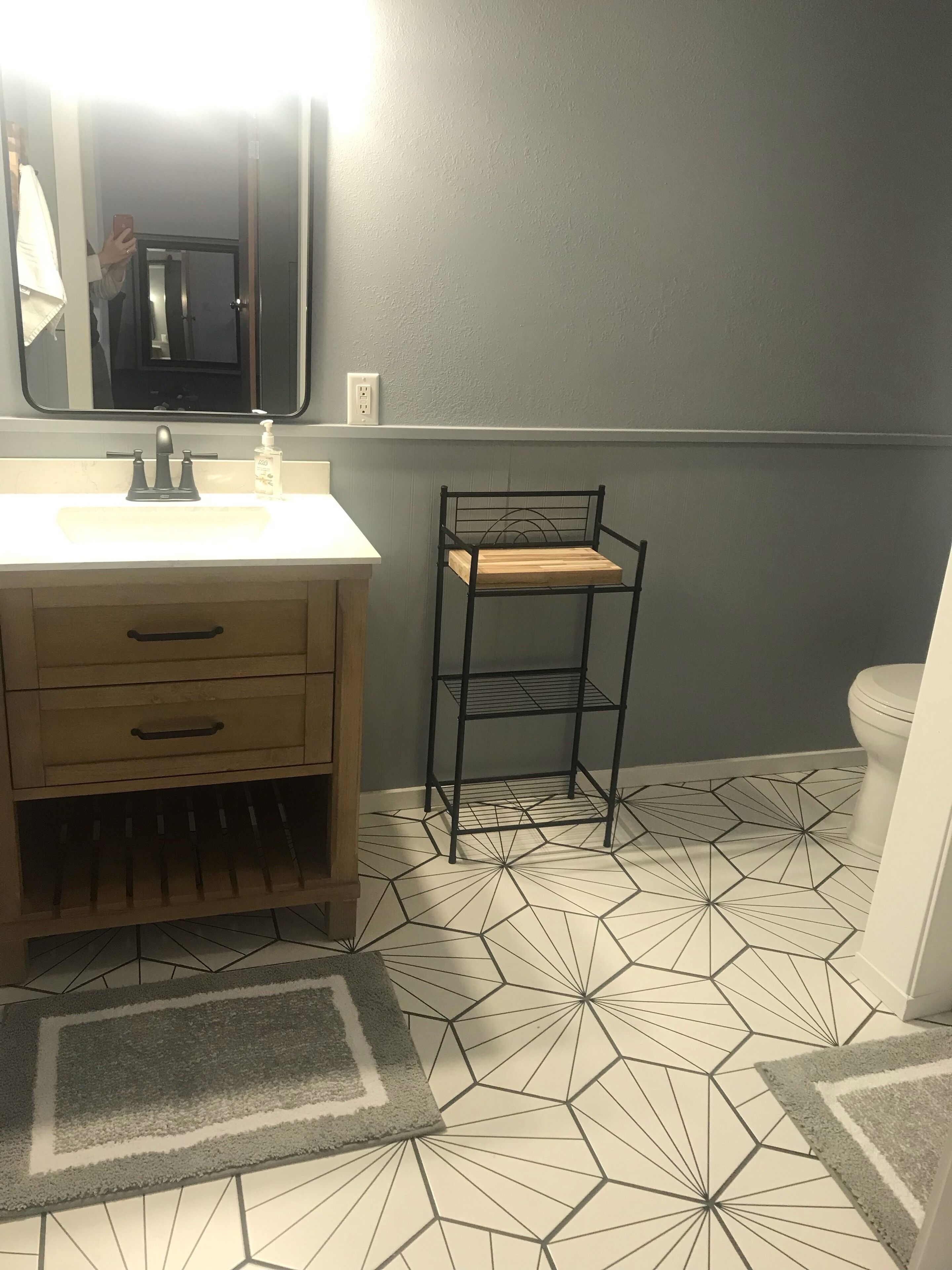 Un baño con un lavabo blanco, un espejo, una silla de metal negro, un inodoro y un suelo con estampado geométrico.