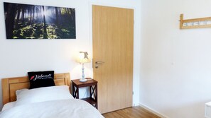 Un dormitorio con cama, una mesita de noche con lámpara, una puerta y un cuadro colgado en la pared que muestra un bosque.