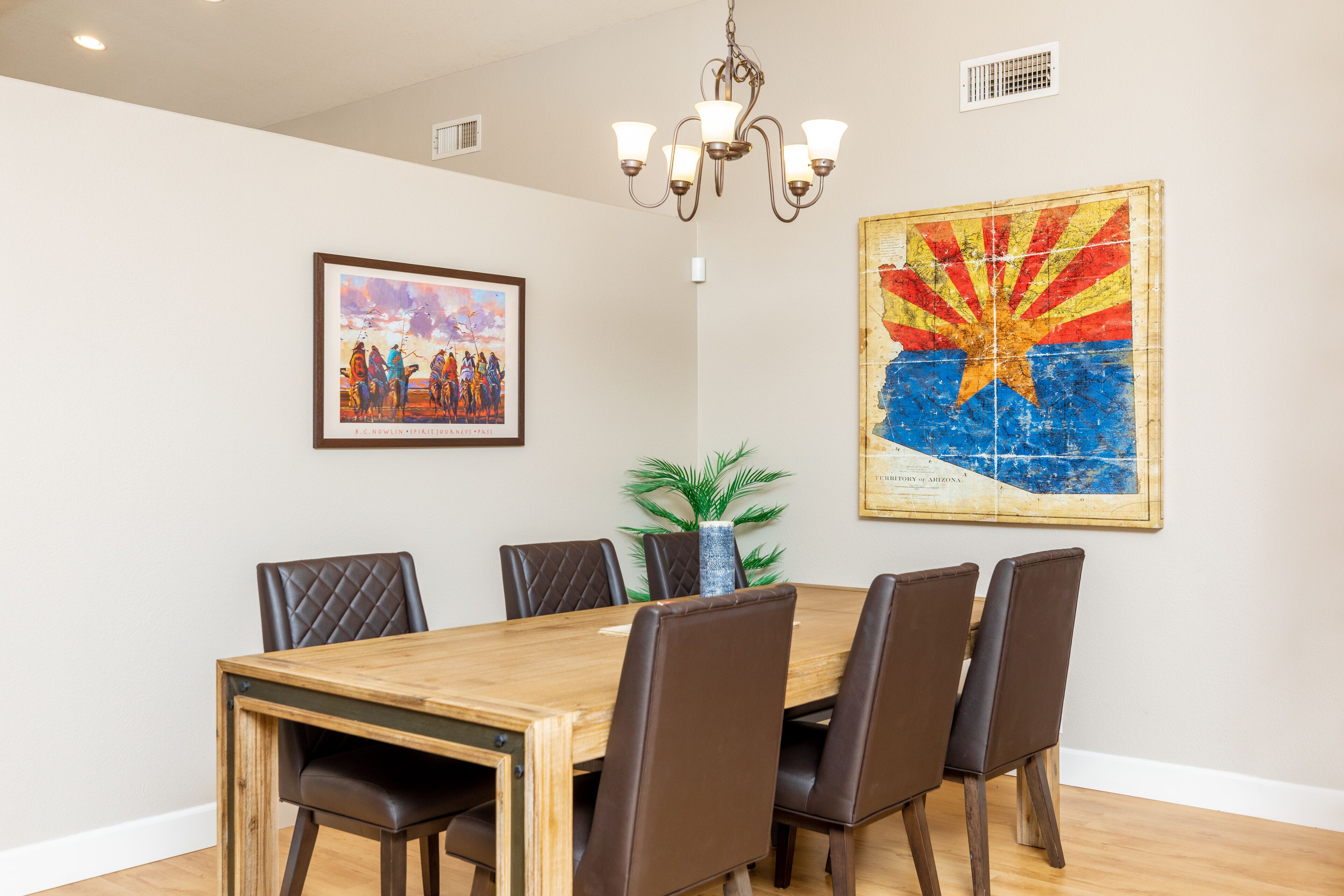A dining area with a wooden table and six chairs, a chandelier, a framed painting, and a map of Arizona on the wall.
