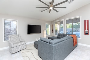 A living room with a grey sectional sofa, a patterned armchair, and a flat-screen TV mounted on the wall.