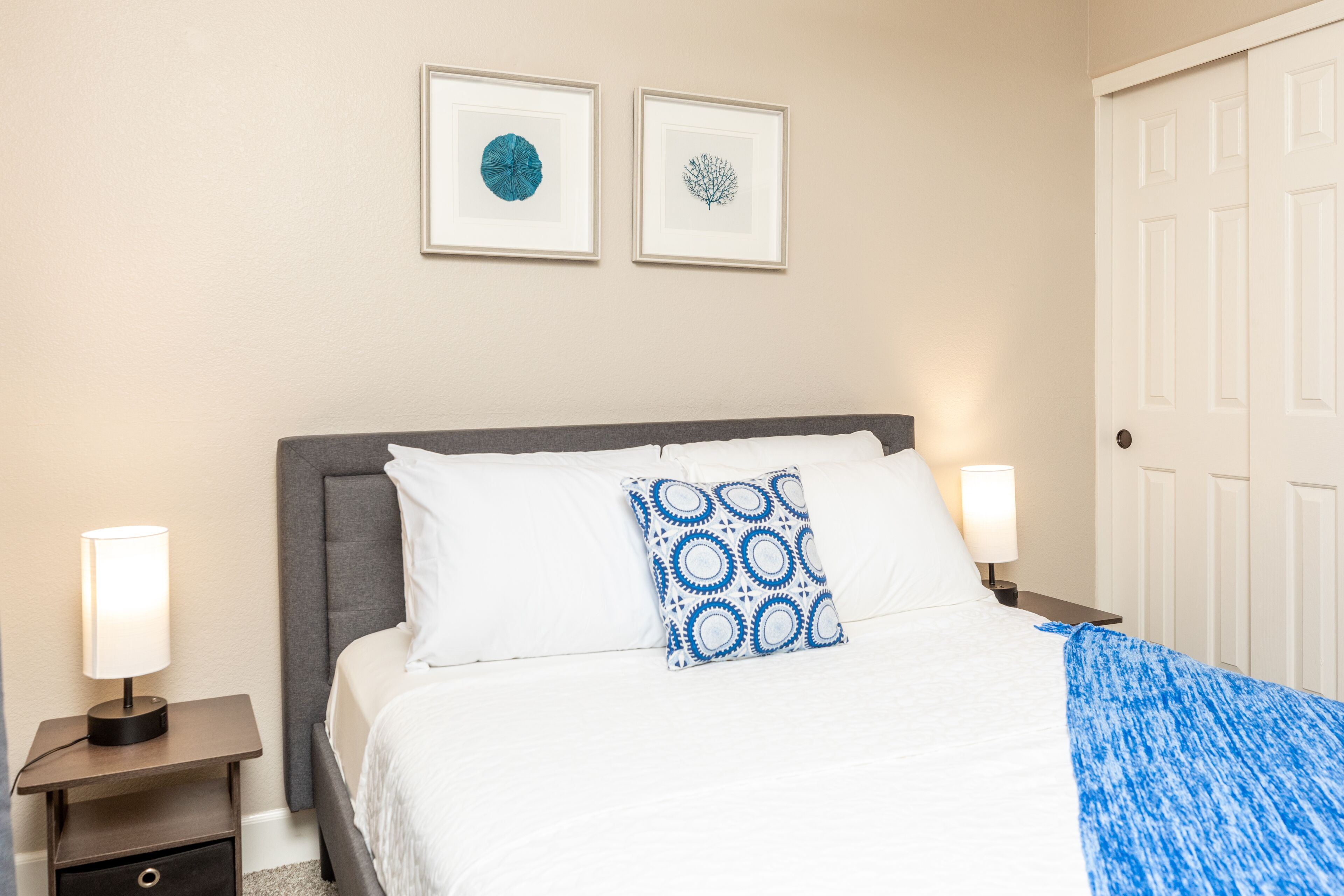 Una cama bien hecha con ropa blanca y una almohada estampada en azul, una mesita de noche con una lámpara, y dos cuadros en la pared.