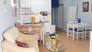 Un soggiorno con un divano beige, un tavolino in vetro con un vaso, e una zona pranzo con sedie bianche e una tovaglia blu.