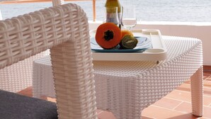 Una sedia e un tavolo in vimini con una bottiglia, un bicchiere e della frutta su una terrazza affacciata sul mare.