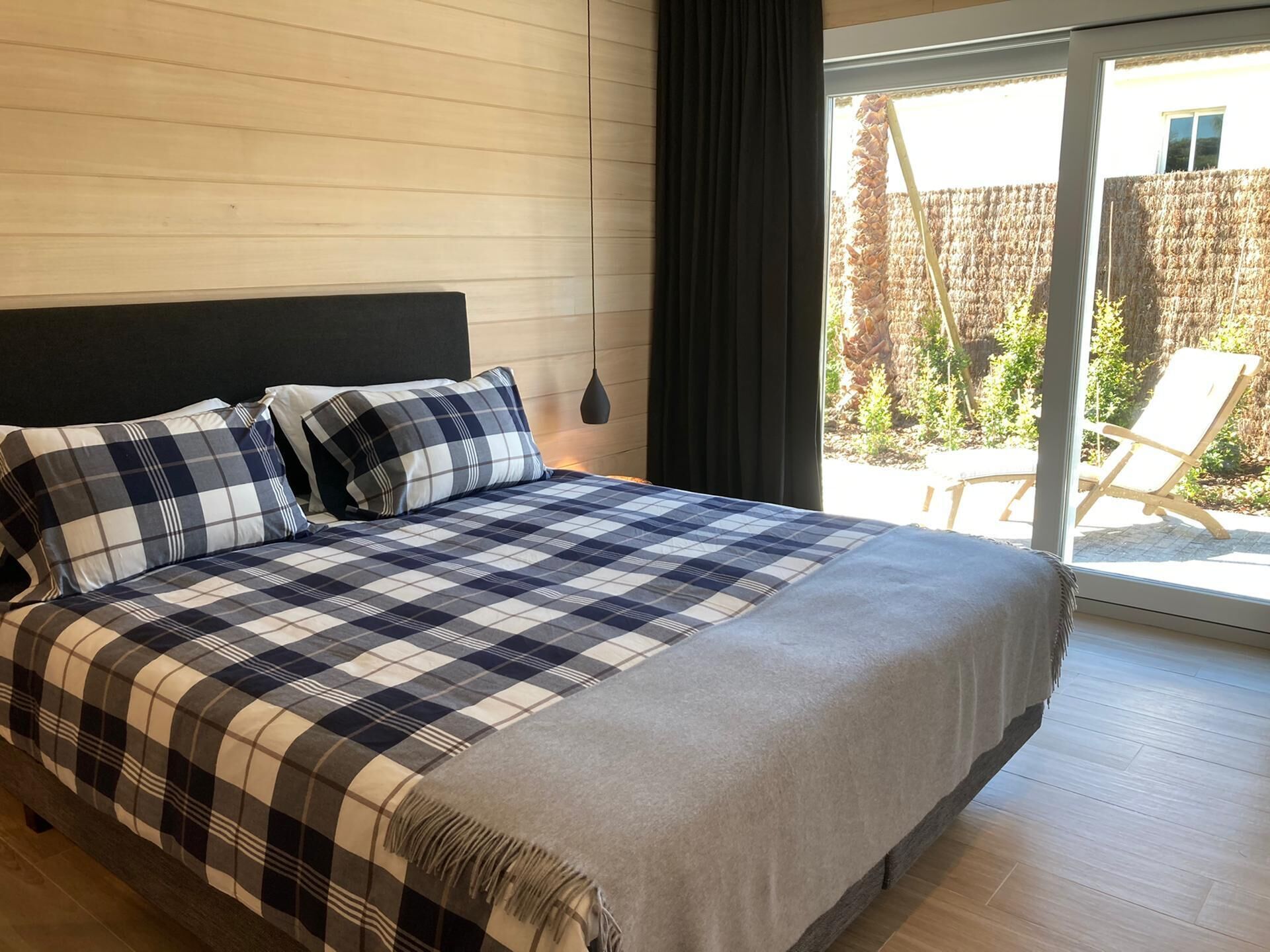 Une chambre à coucher avec un grand lit, une tête de lit sombre et une vue sur un jardin, accessible par une porte-fenêtre coulissante.