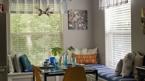 Un coin repas comprenant une table, des chaises et un banc. On y trouve également un ventilateur de plafond, une fenêtre avec des stores et un tableau accroché au mur.