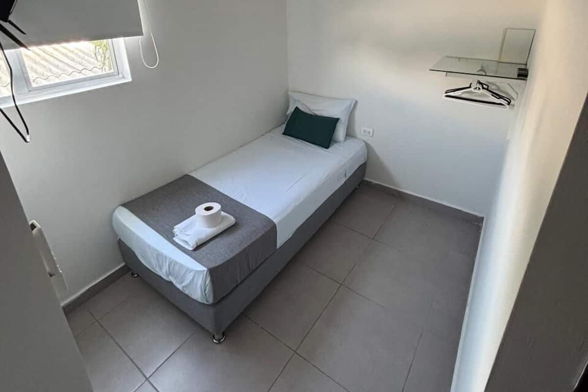 A small, compact room with a single bed, a window, a mirror, and a wall-mounted air conditioner.