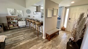 A modern kitchen with a central island, bar stools, and a bench. There is a dining area with a wooden table and chairs. The kitchen has a clock on the wall and a vase with dried plants. - Relaxing desert retreat with amazing mountain and lake view on the 7th fairway! (Palm Desert)