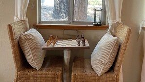 A cozy corner with two wicker chairs, a small table with a chessboard, and a window with sheer curtains.
