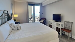 Een hotelkamer met een groot bed, een bureau met een televisie, een stoel, een plafondventilator en uitzicht op de oceaan door de schuifdeuren.
