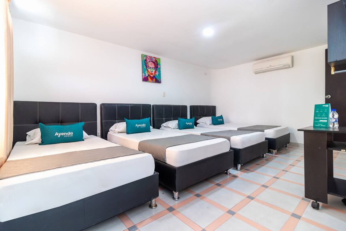 A hotel room with three beds, each with a black headboard, white bedding, and teal pillows labeled "Ayenda Hotelier."