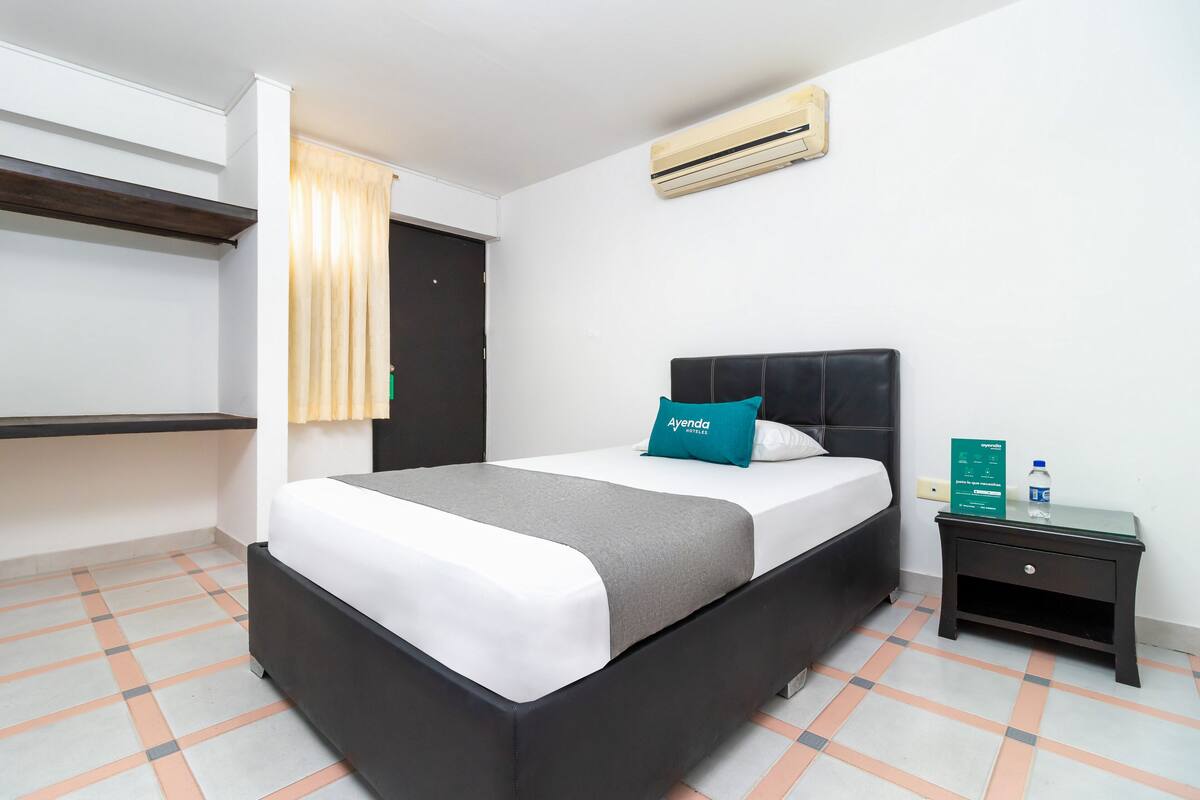 A neatly arranged hotel room with a bed, a nightstand, a wall-mounted air conditioner, and a check-in desk.