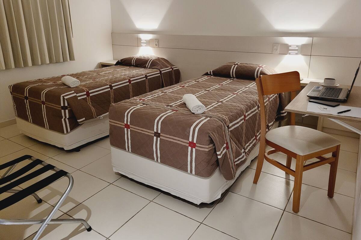 Habitación de hotel con dos camas, un escritorio con una computadora portátil, una silla y una ventana con persianas.