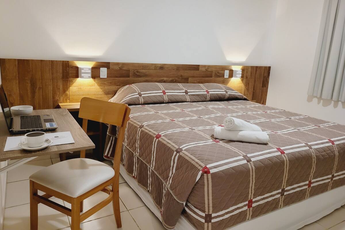 Habitación de hotel ordenada con cabecera de madera, cama con colcha acolchada, escritorio con una computadora portátil, una silla y una mesita de noche con una taza y un teléfono.