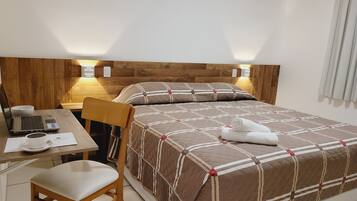Quarto de hotel bem organizado, com cabeceira de madeira, cama com colcha acolchoada, escrivaninha com um laptop, uma cadeira e mesa de cabeceira com uma xícara e um telefone.