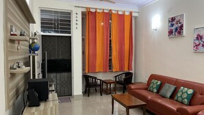 A living room with a brown leather sofa, a wooden coffee table, and a dining area with a table and chairs. There is a globe on a shelf, a television on a stand, and a window with orange curtains.