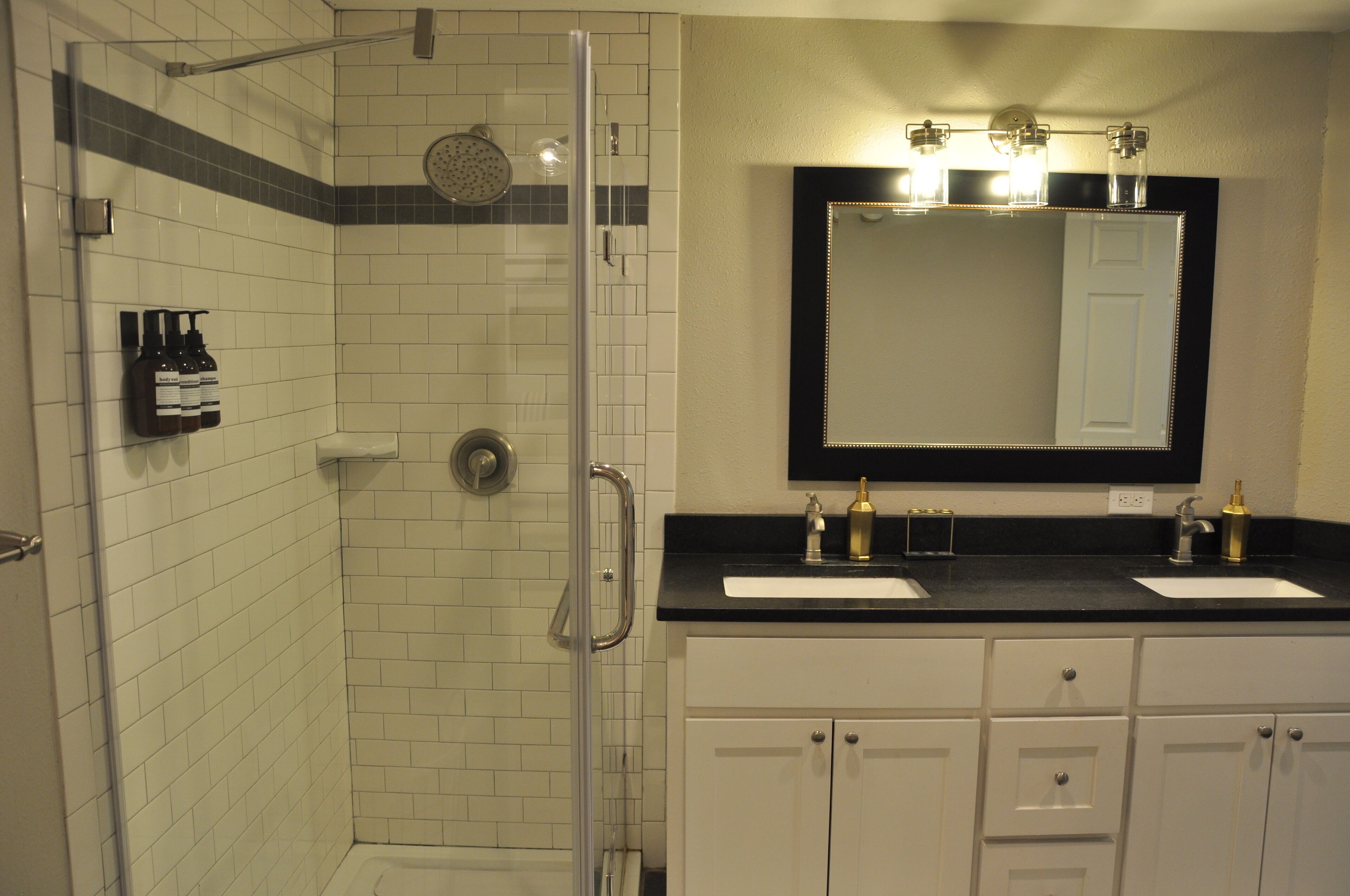 A bathroom with a glass-enclosed shower, two sinks, and a large mirror with lights above.